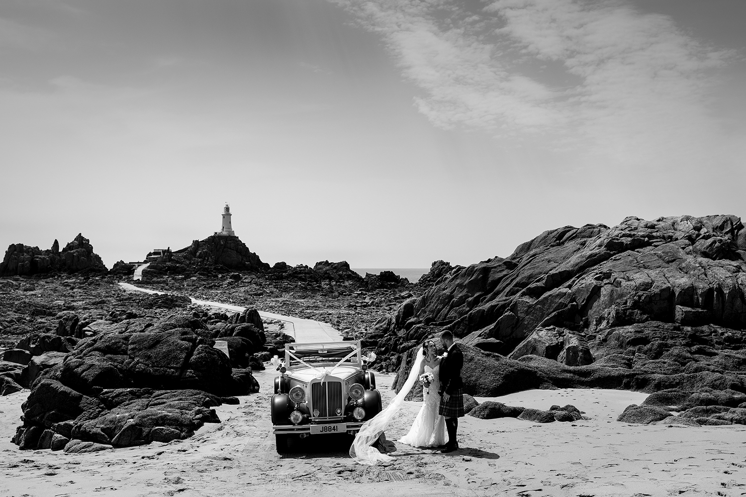 The newly wed couple embrace near their vintage bridal car with the the iconic Corbiere Lighthouse Jersey Channel Islands, in the distance